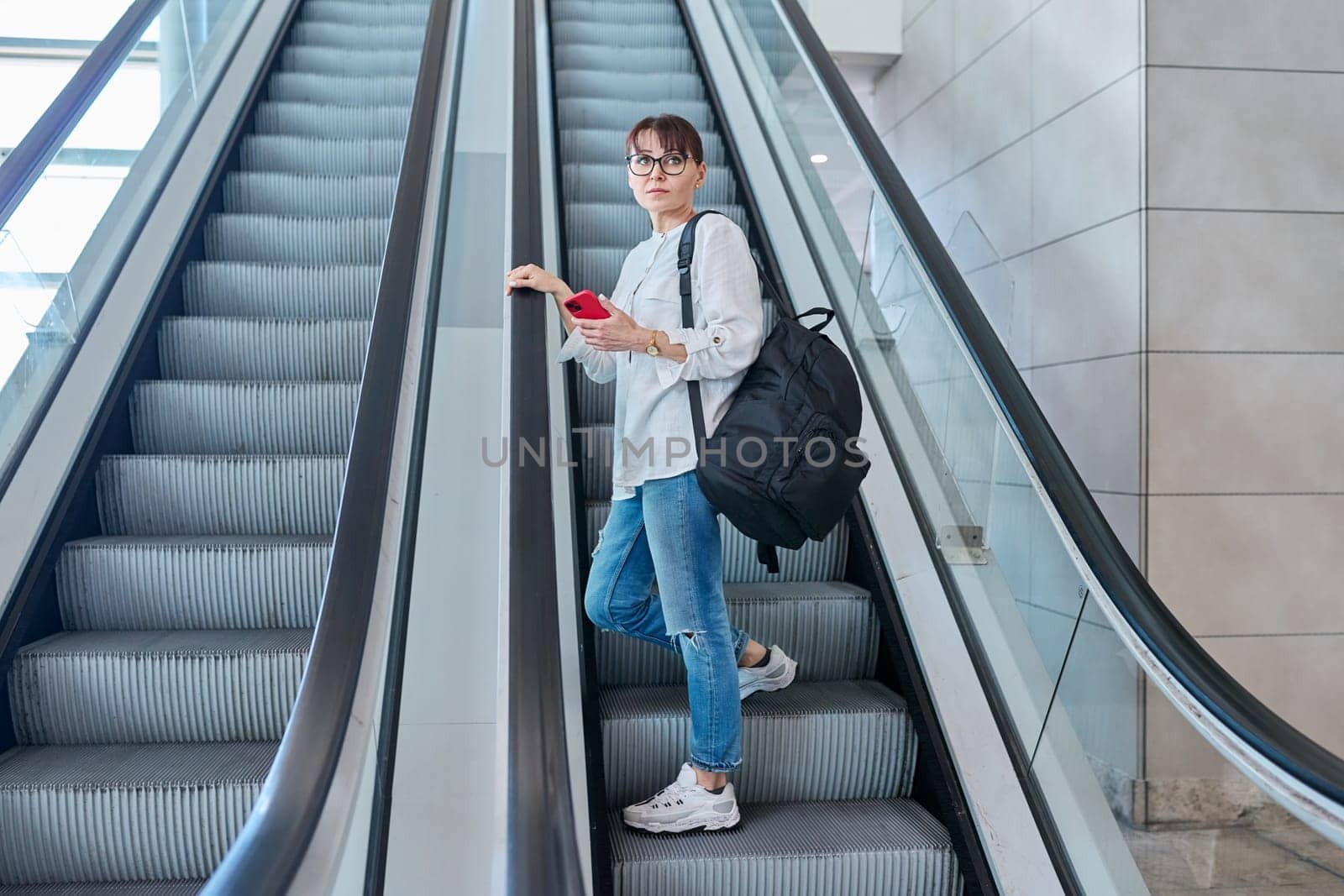 Middle-aged woman with backpack in casual style on escalator by VH-studio