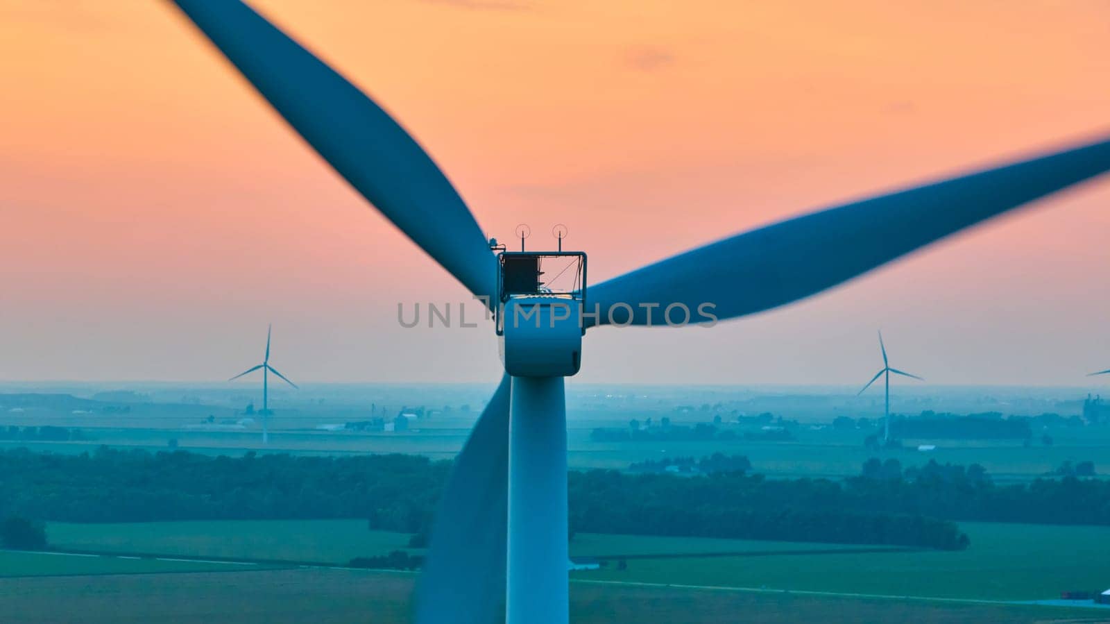 Image of Aerial straight of back of wind turbine with distant turbines in green farmland at sunset