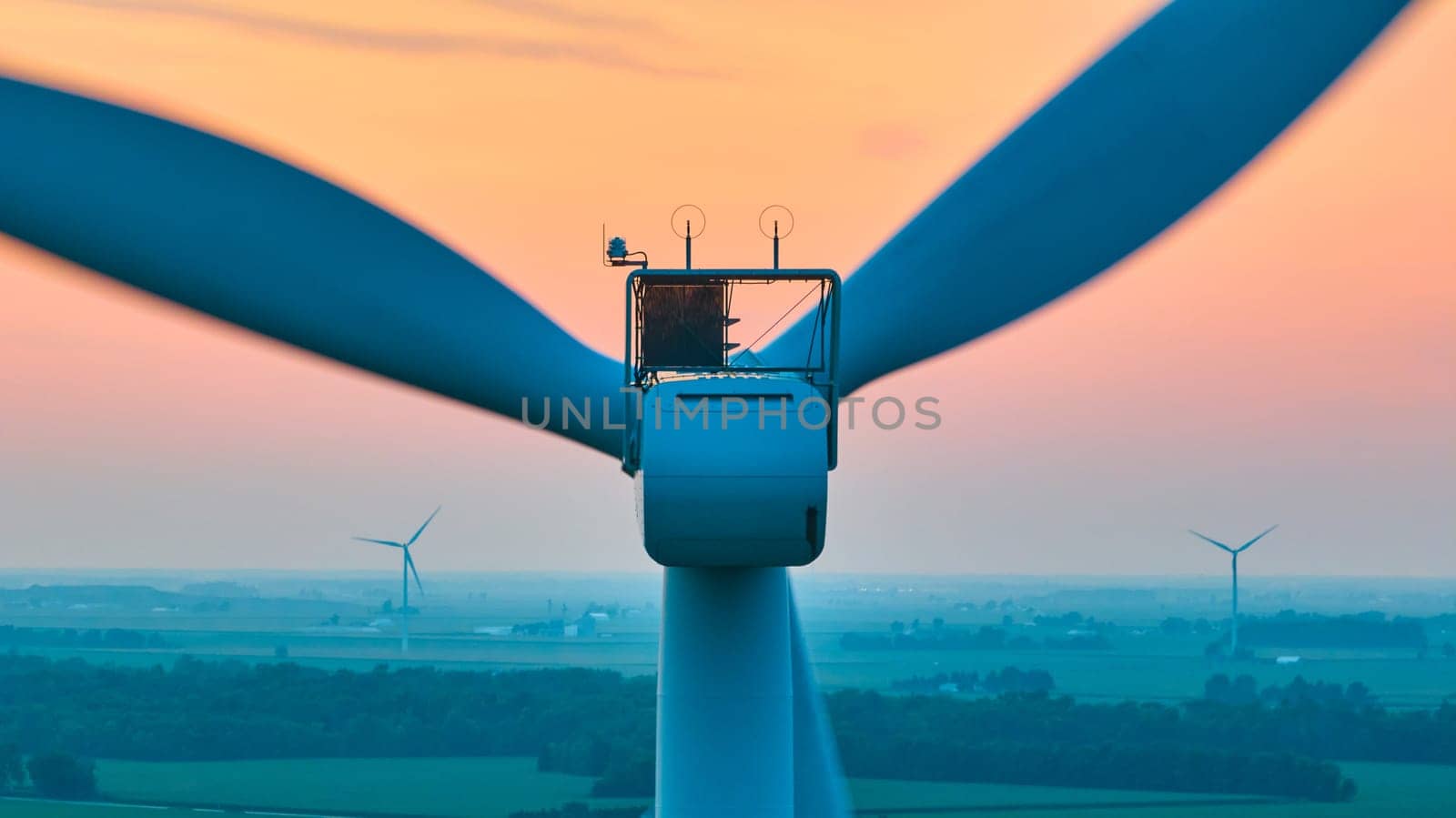 Image of Close up and straight on aerial of wind turbine with distant turbines in green farmland at sunset
