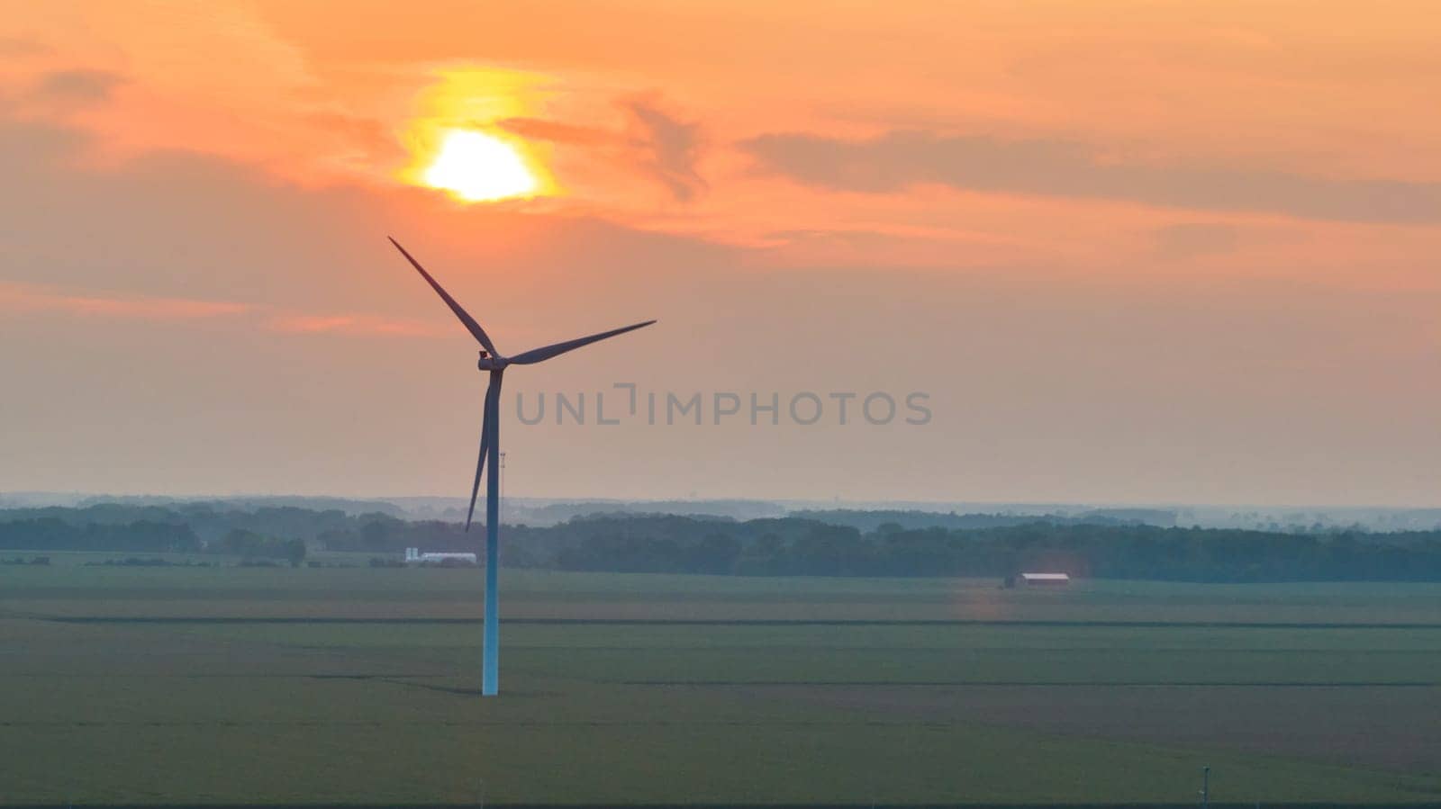 Lone wind turbine in field with orange sun over it at sunset aerial by njproductions