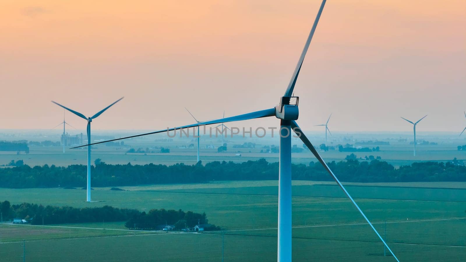 Image of Aerial crisp wind turbine at sunset with orange sky with wind farm behind it and green fields below