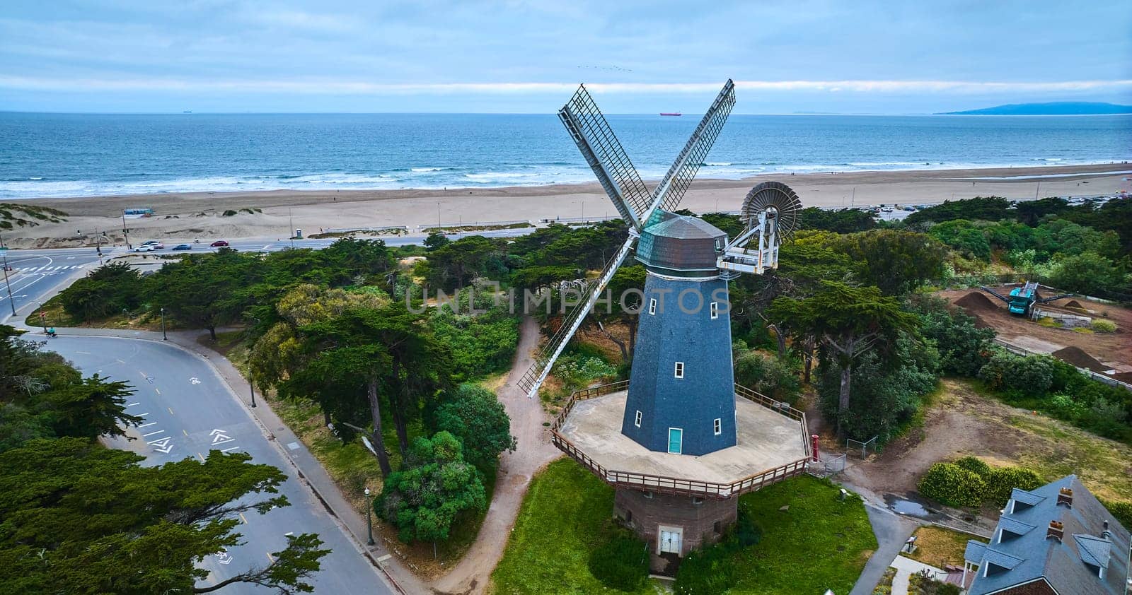 Image of Blue murphy windmill surrounded by trees and roads overlooking blue ocean with sandy shore aerial