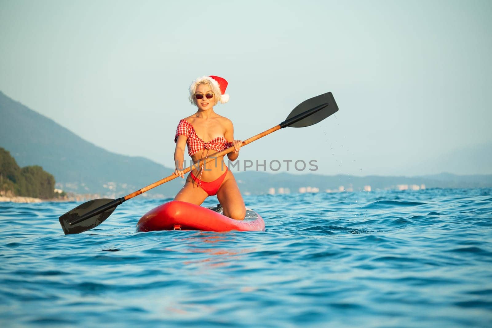 Sexy girl in a Santa Claus hat floats on the sea on a Sup Board in a red bikini celebrating Christmas at a resort in a hot country by Rotozey
