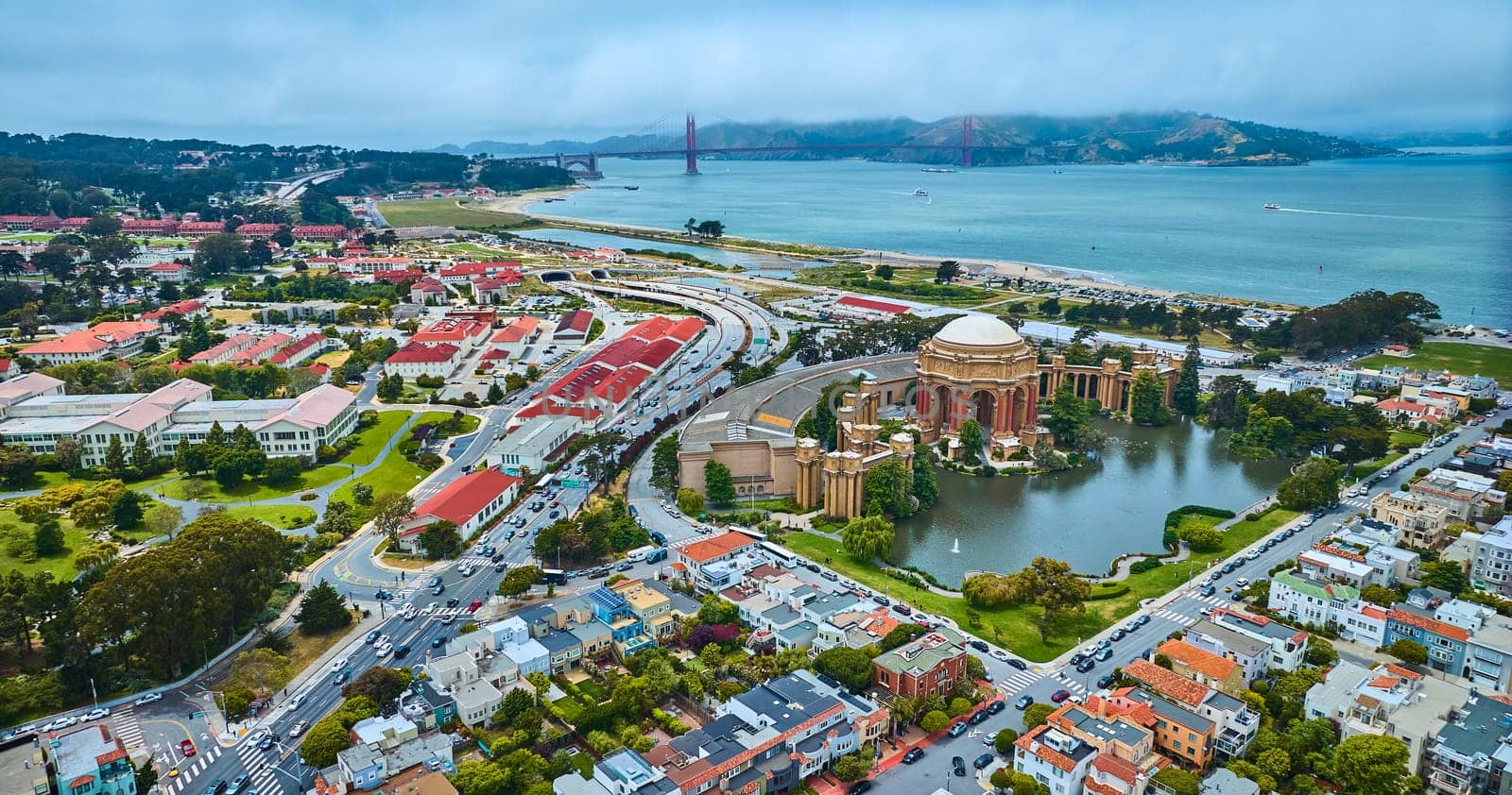 Image of Palace of Fine Arts rotunda and colonnade around pond with Golden Gate Bridge in distance aerial
