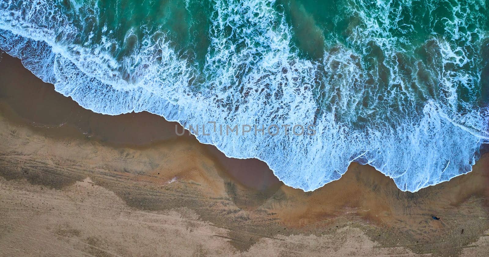 Image of Downward aerial of receding green ocean waves on top with dark tan sandy beach on bottom