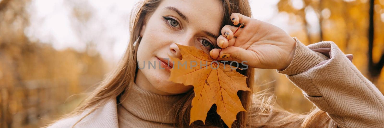 Portrait of a woman with an autumn maple leaf. Railway, autumn leaves, a young long-haired woman in a light coat coat, close-up.