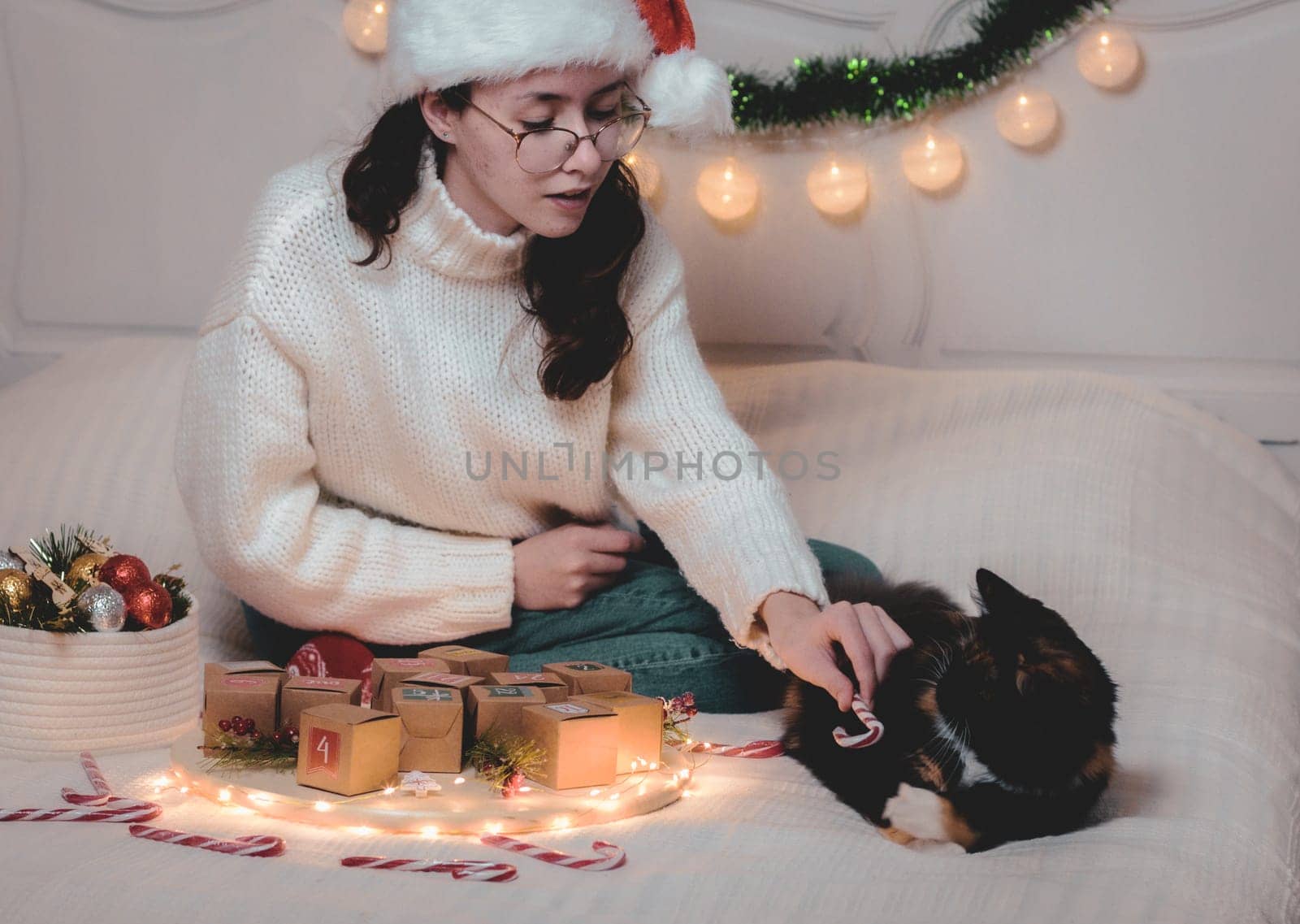 Beautiful caucasian teen girl in santa claus hat plays with purebred cat,sitting on bed with advent calendar and christmas decorations in bedroom,closeup side view.