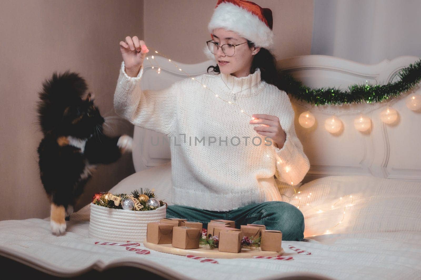 Beautiful caucasian teenage girl in santa claus hat plays with purebred cat with burning garland while sitting on bed with advent calendar and christmas decorations in bedroom, closeup side view.