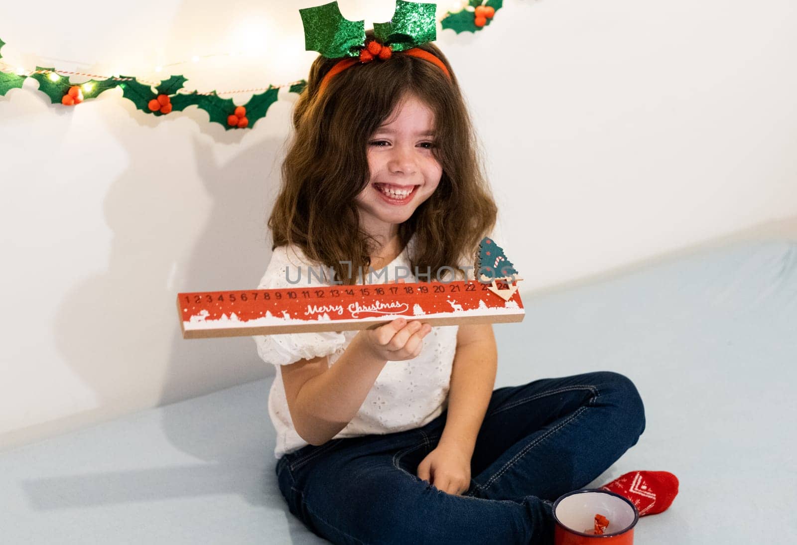 Beautiful little caucasian girl with a happy smile on her face holds a ruler of the advent calendar moved the Christmas tree to the number 24 in her hands, sits on the bed, looking to the side, close-up view.