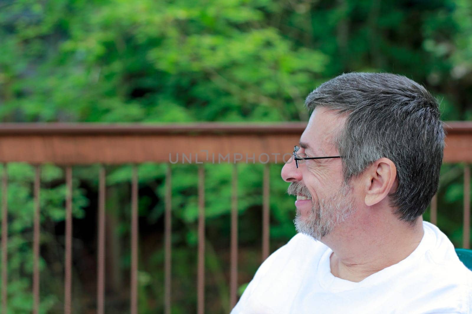 Profile view of a mature man smiling outdoors with a background of a deck railing and trees.