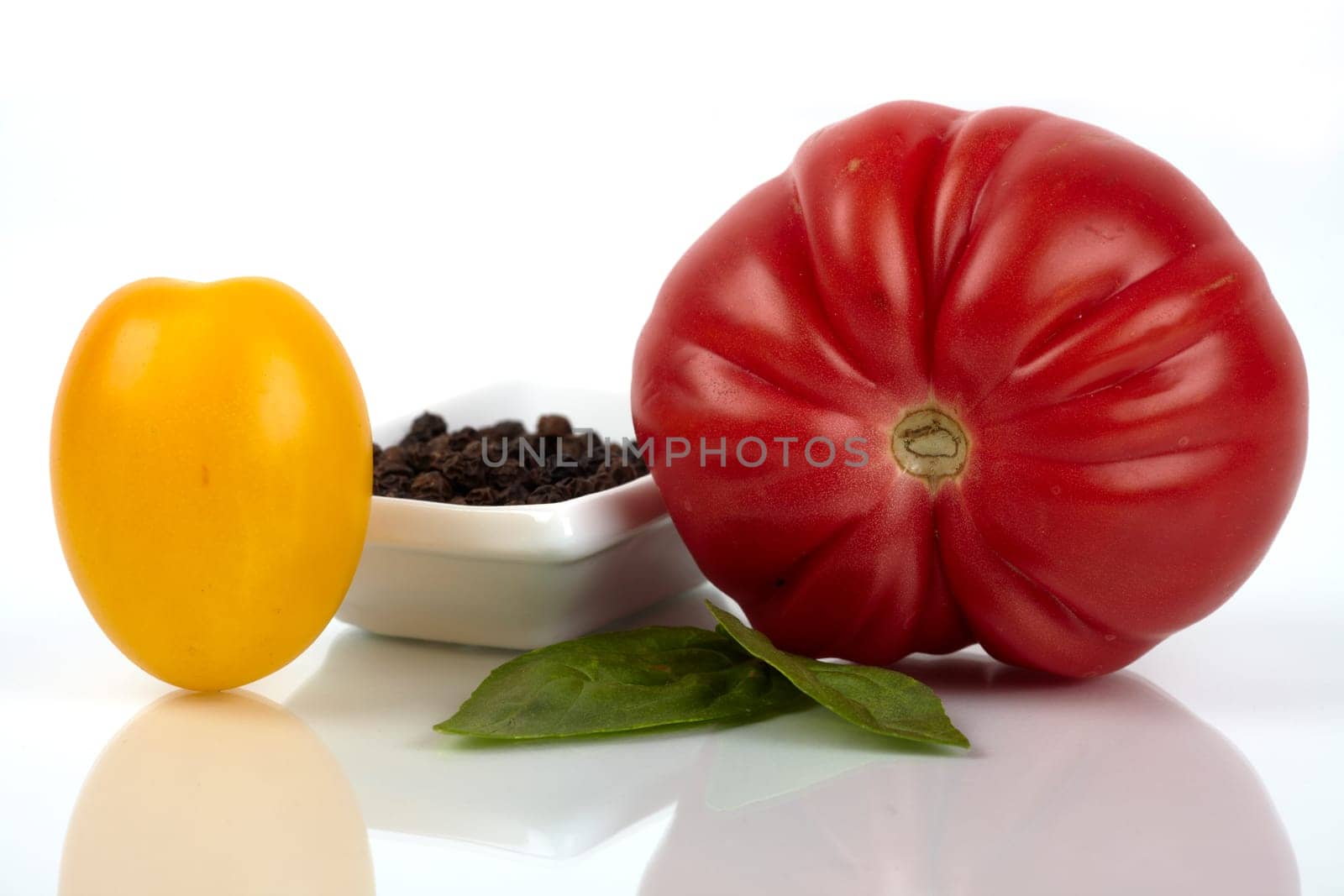 Two different organic tomatoes with small peppercorns skin and basil leaves,close-up mirrored on glass plate. Two different organic tomatoes with a small cup peppercorns and basil leaves on plate glass mirror closeup.