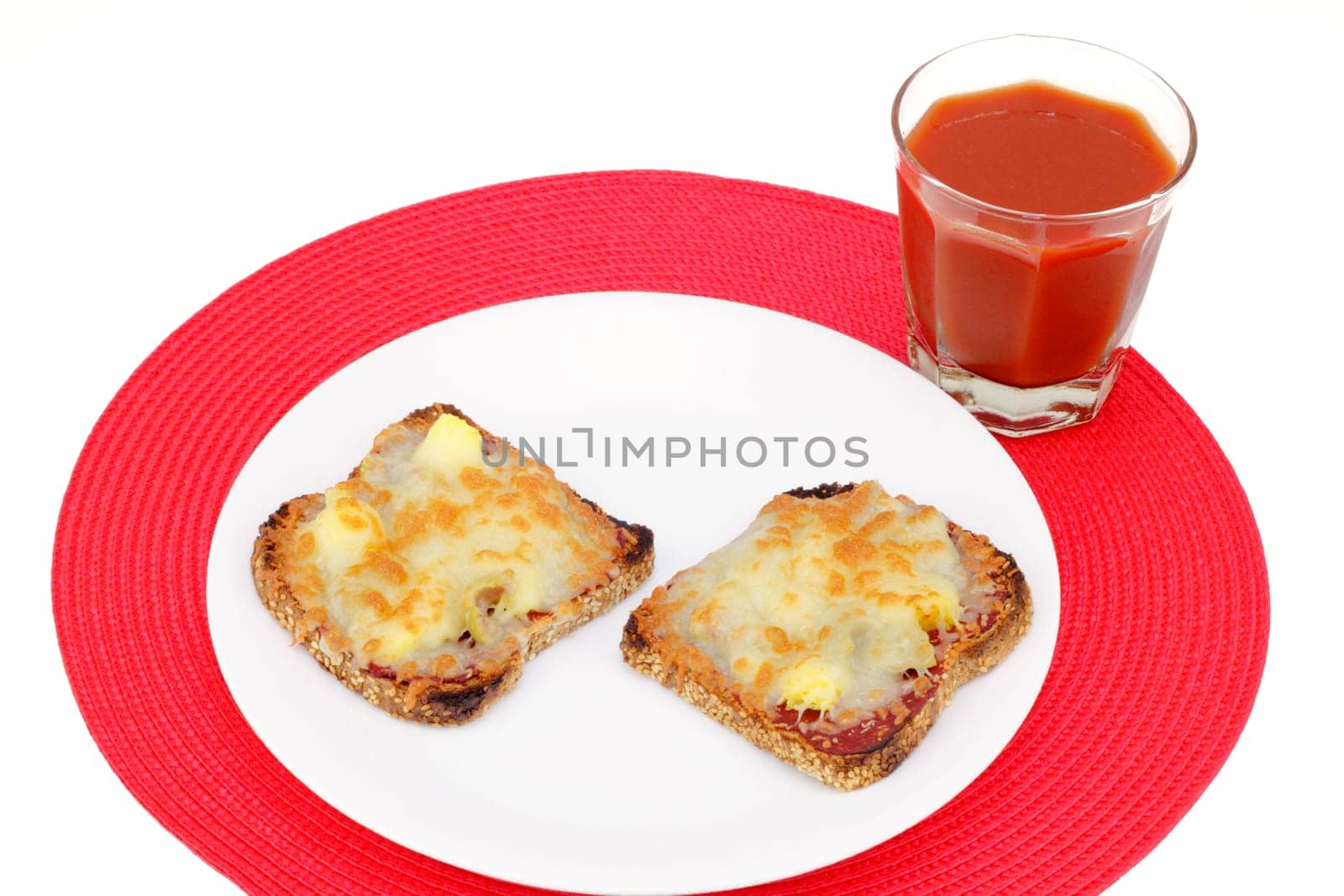 Two healthy slices of sprouted grain bread topped with pizza toppings on a white plate and one glass of vegetable tomato juice on a round red place mat.