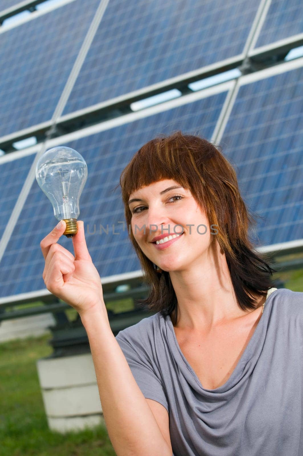 Head-and-shoulder shot of a young woman with a light bulb in her hand in front of a photovoltaic system, looking friendly at the camera.