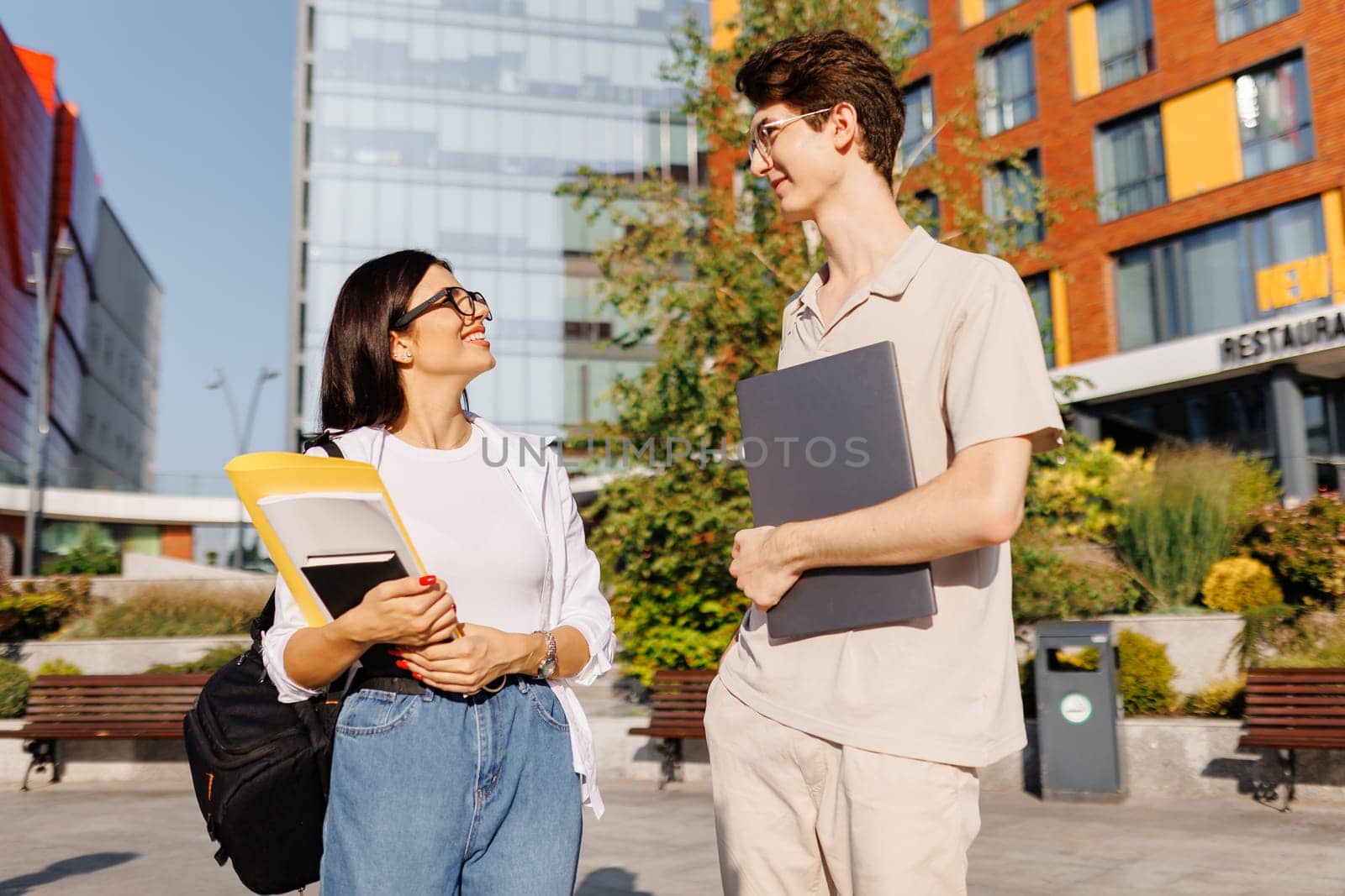 Smiling young students male with laptop and female with books looking at each other while speaking and walking with backpacks on campus street in daylight against blurred green park blue sky buildings