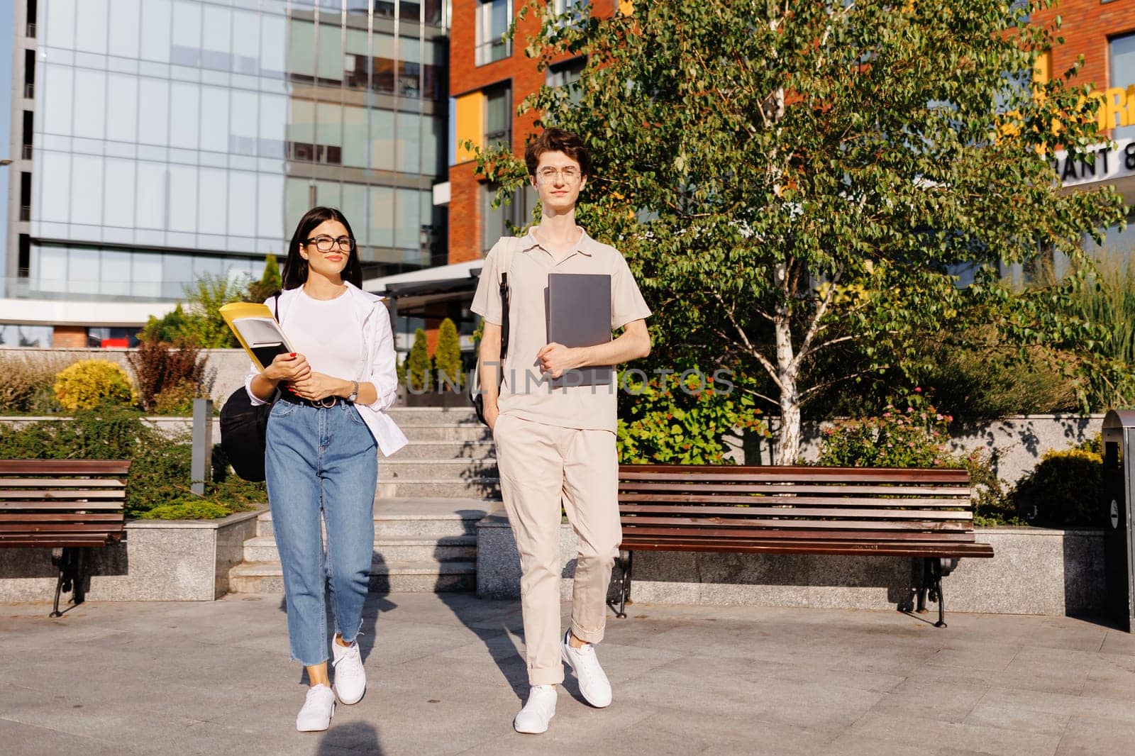 Smiling young male with laptop and female with books looking at camera while standing together on campus street near green trees against blurred college building in daylight