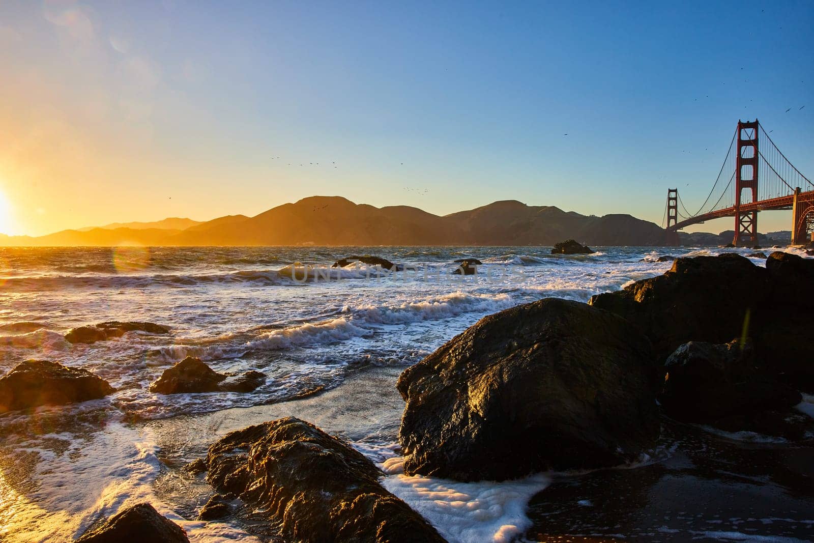 Image of Gorgeous sunset over waves and boulders with Golden Gate Bridge and distant mountains