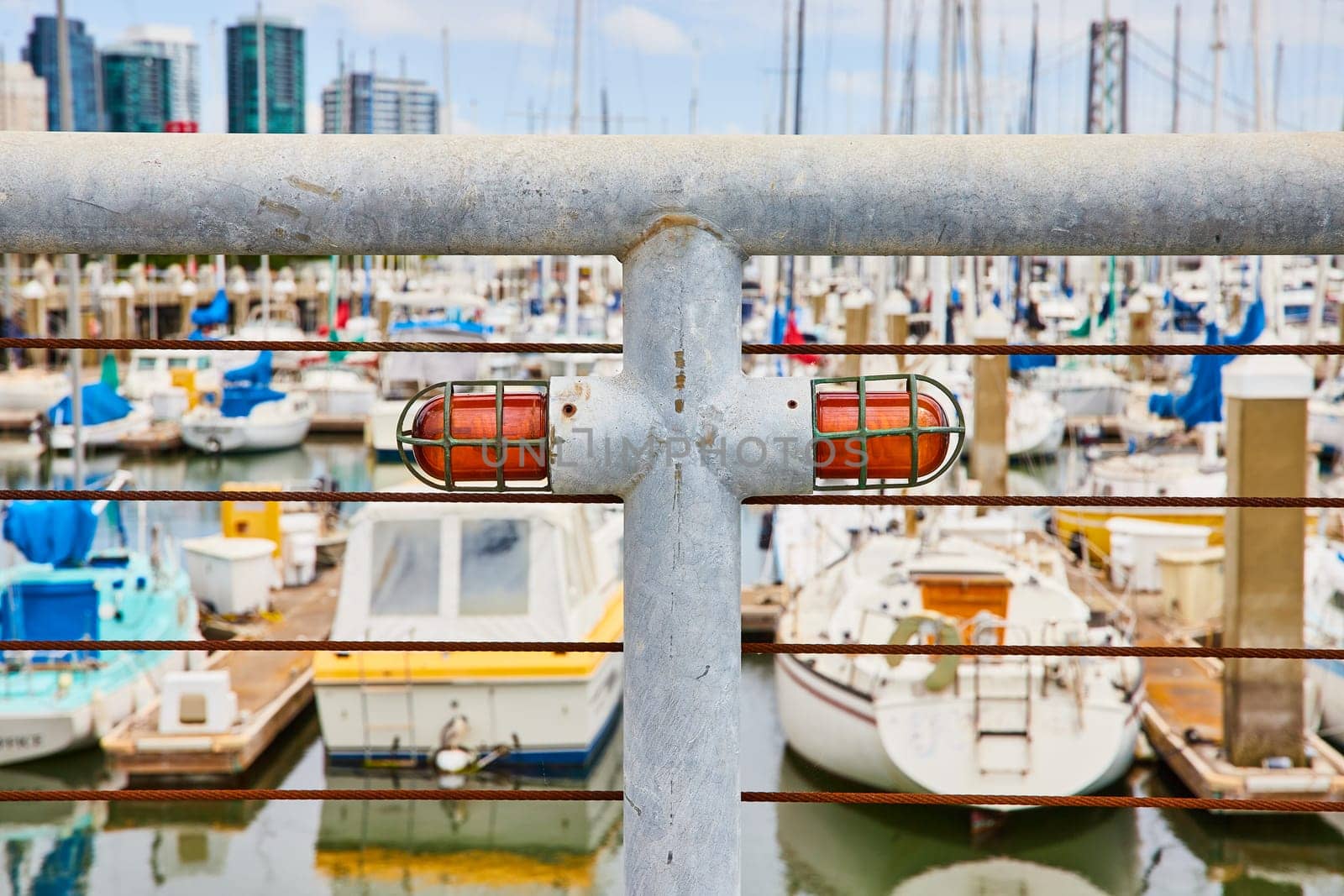 Image of White metal railing with brown iron wires and big heavy duty red lights with blurry boats and city