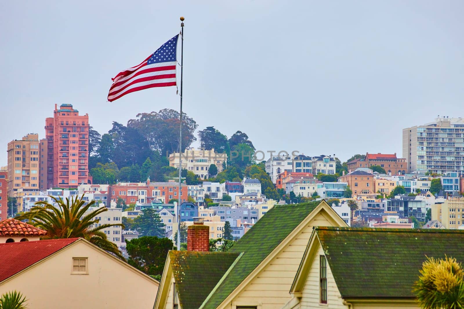 Image of American Flag waving in wind over green residential rooftops with overcast sky and city behind it
