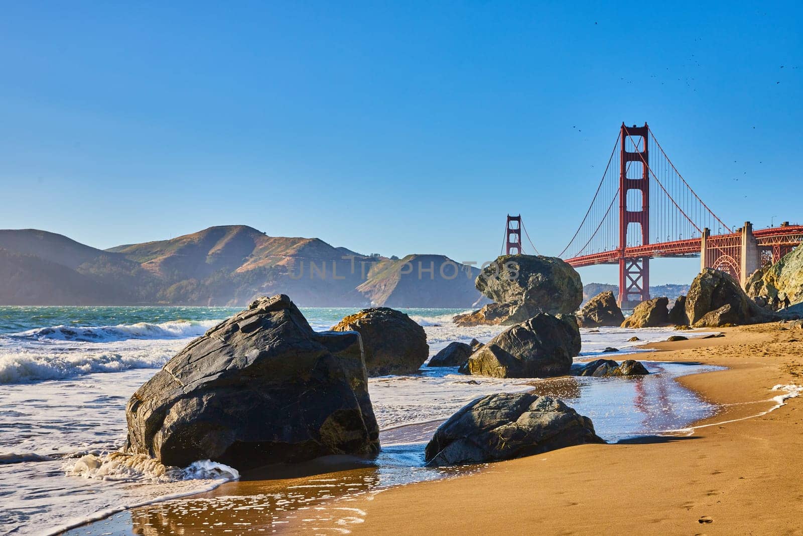 Image of Sunny beach with waves crashing against boulders and distant Golden Gate Bridge
