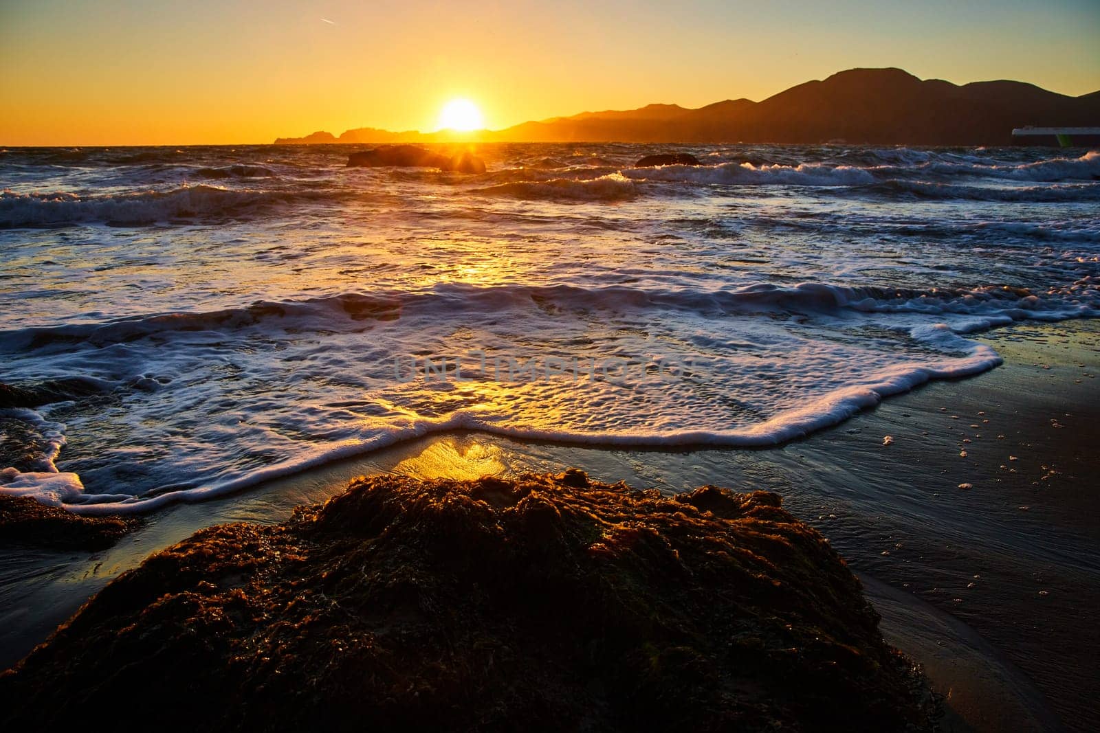 Image of Gorgeous sun setting over distant mountains with waves crashing against the shore