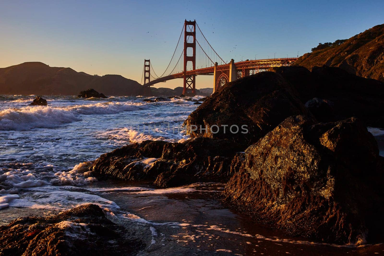 Image of Golden light striking dark mossy rocks with breaking wave and distant Golden Gate Bridge