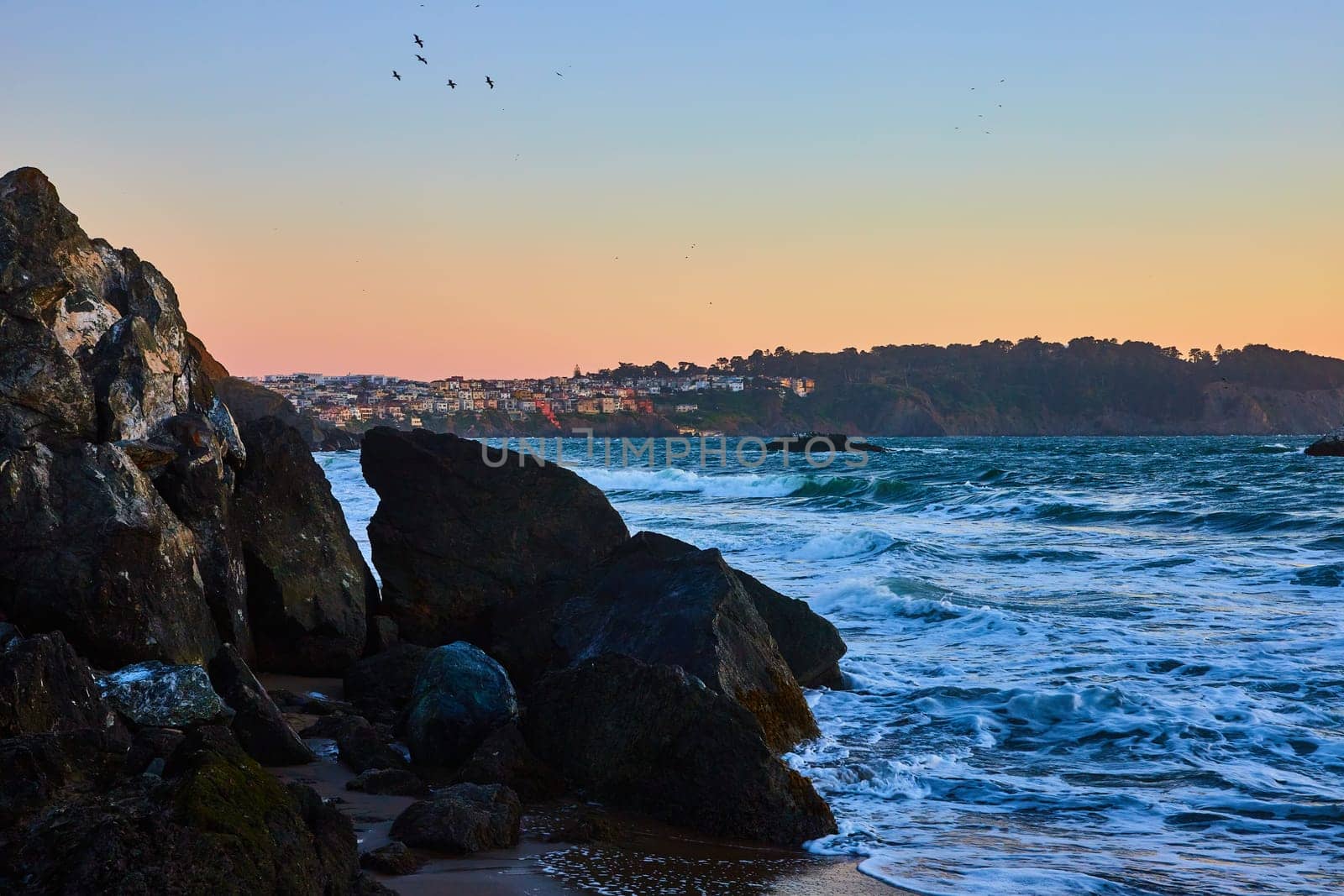 Image of Blue hour on sandy shore with seafoam from waves around boulders and city above cliff