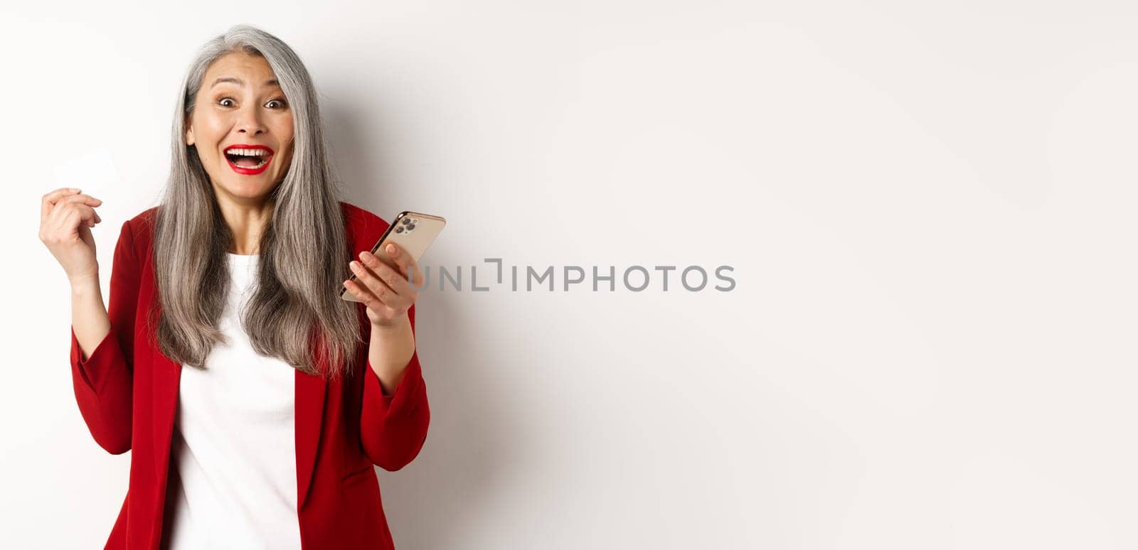 Excited asian businesswoman in red blazer, holding plastic credit card and smartphone, smiling happy at camera, standing over white background.