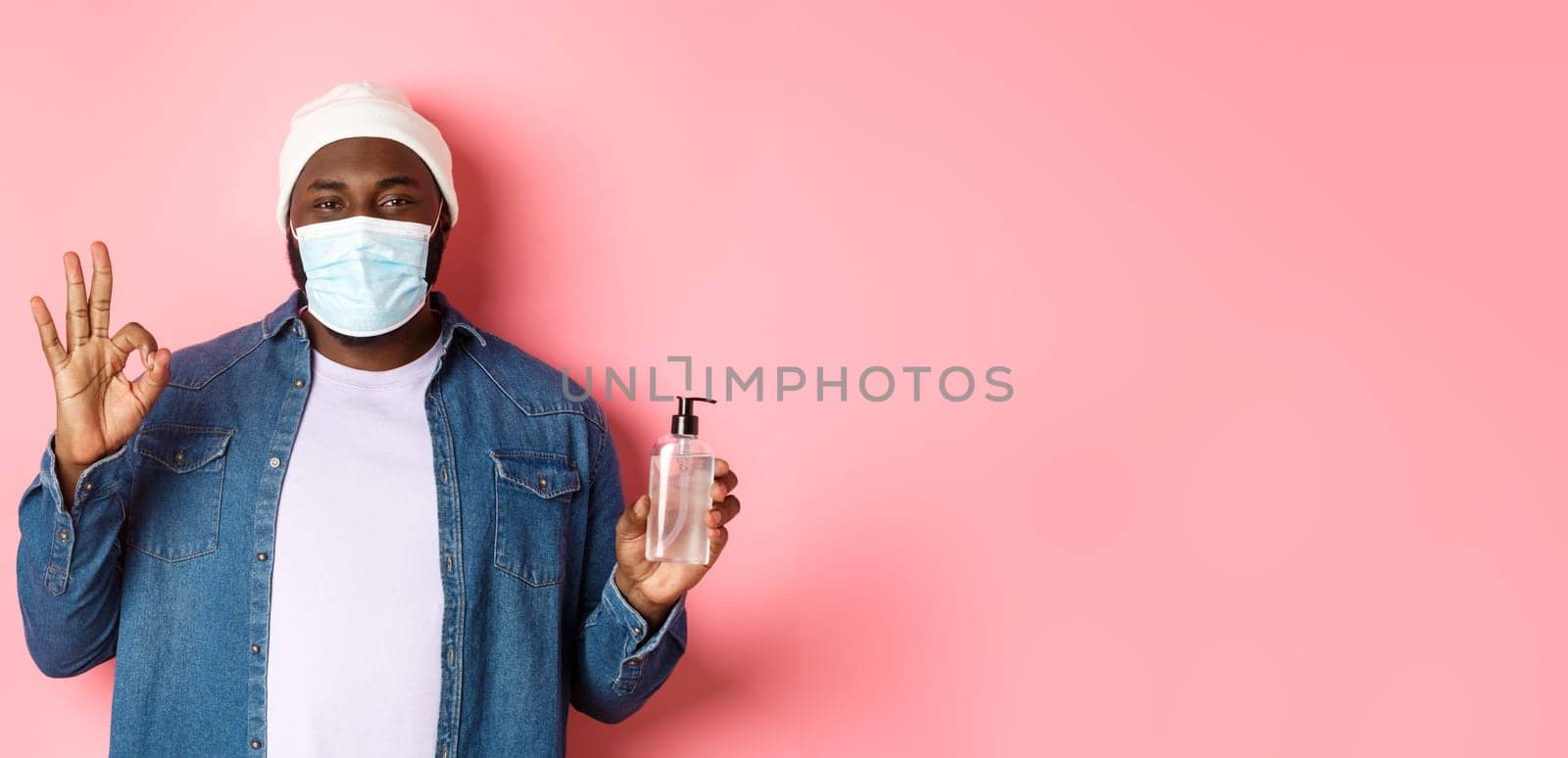 Covid-19, lifestyle and lockdown concept. Handsome african-american hipster guy in face mask and beanie, showing hand sanitizer and okay sign, advice to use antiseptic.