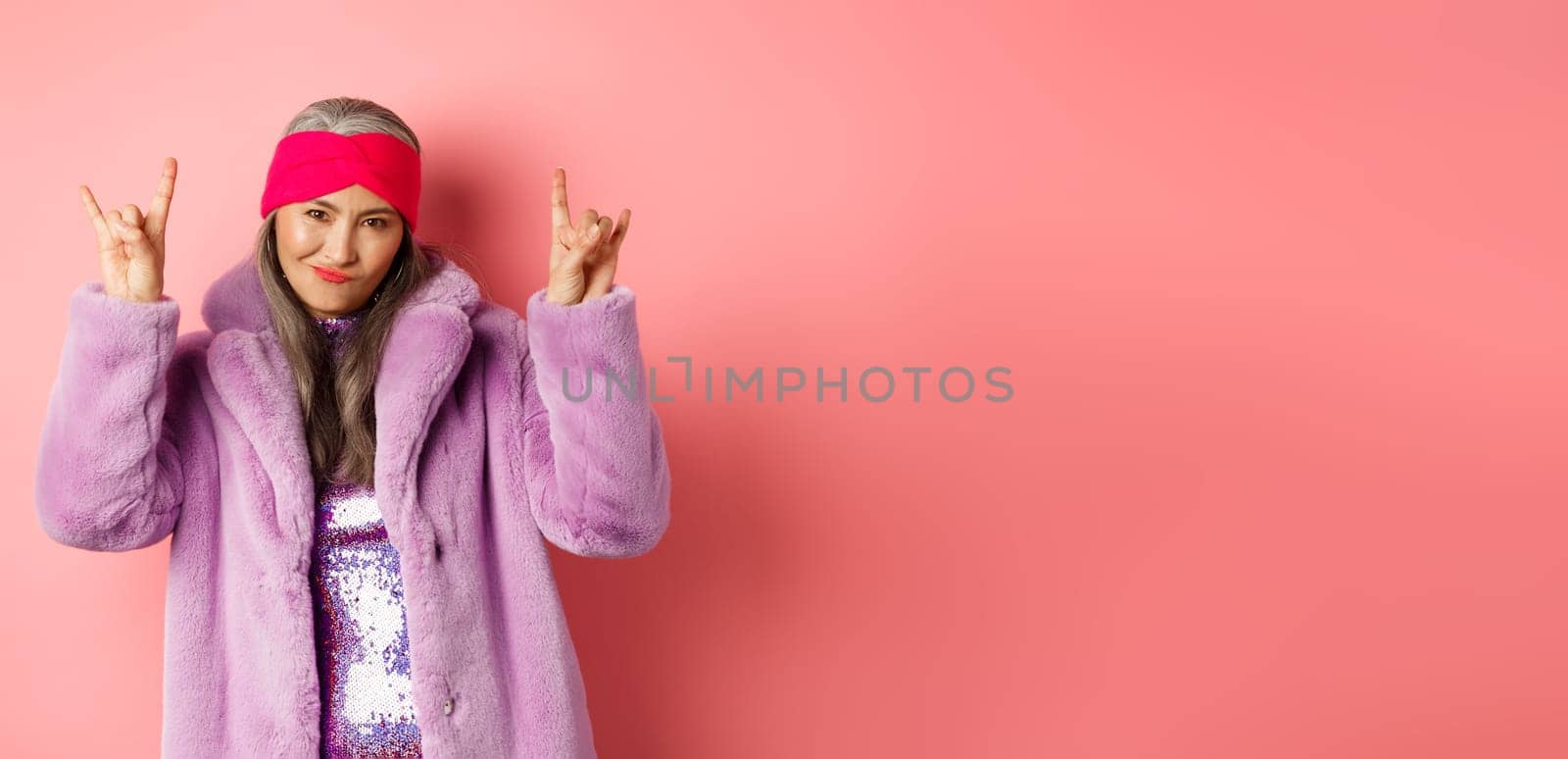 Cool asian mature woman in fashionable faux fur coat, showing rock-n-roll horns signs and smiling sassy at camera, having fun, standing over pink background.