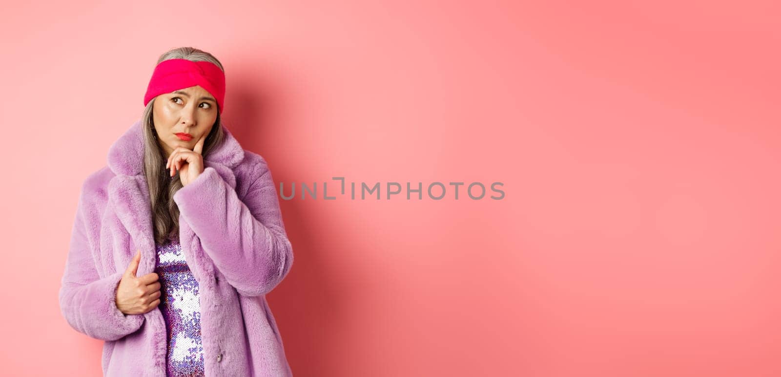 Fashionable asian senior woman looking doubtful, thinking and frowning indecisive, standing in purple faux fur coat and looking left, standing over pink background.