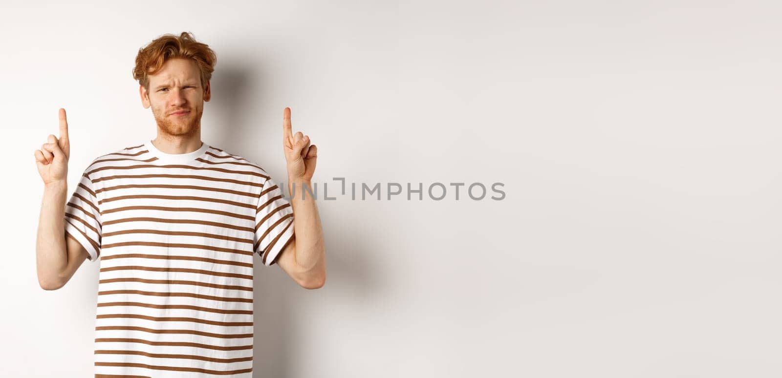 Skeptical handsome redhead man pointing fingers up, grimacing and looking doubtful at camera, standing indecisive over white background.