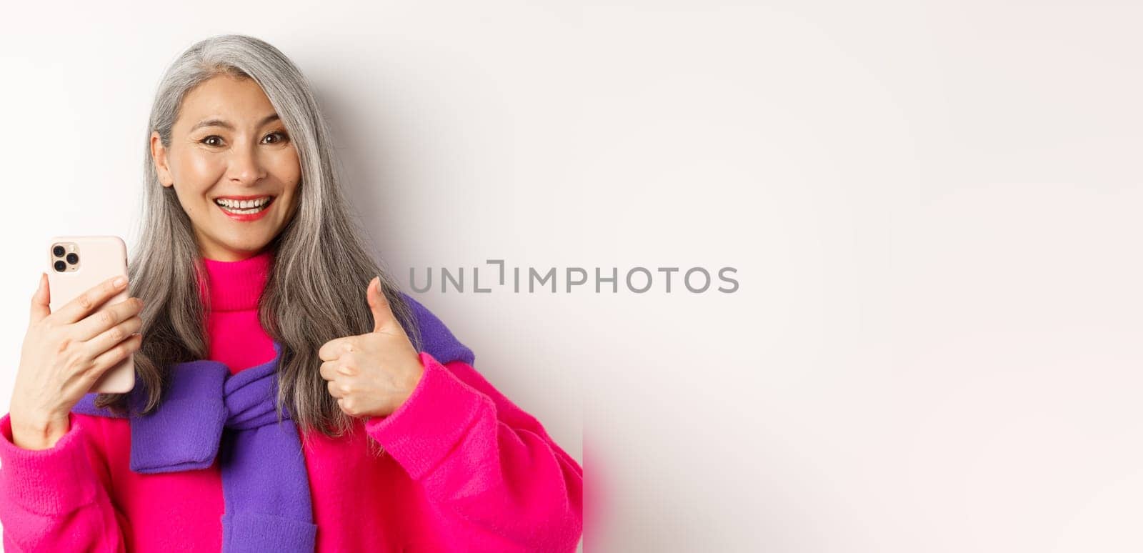 Online shopping. Close up of fashionable asian senior woman showing thumbs-up, using smartphone and approve something, standing over white background.
