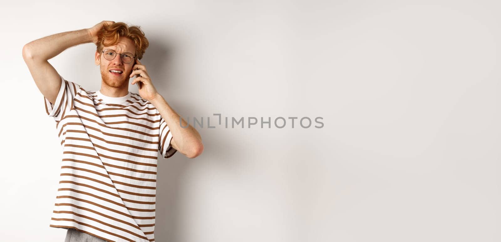 Puzzled redhead guy talking on phone, scratching head and looking confused or indecisive, standing over white background by Benzoix