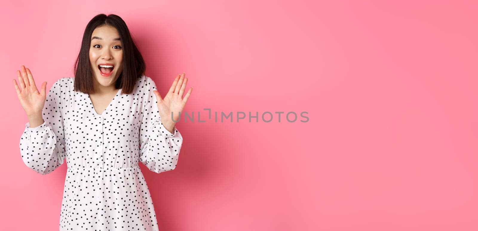 Happy and surprised asian girl receive surprise, looking glad hearing good news, standing amazed in dress against pink background.