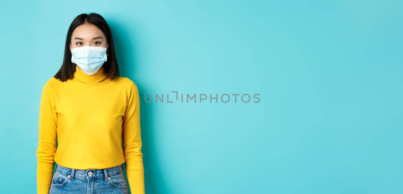 Covid-19, social distancing and pandemic concept. Teenage asian woman in medical mask, looking at camera and standing against blue background in yellow sweater.