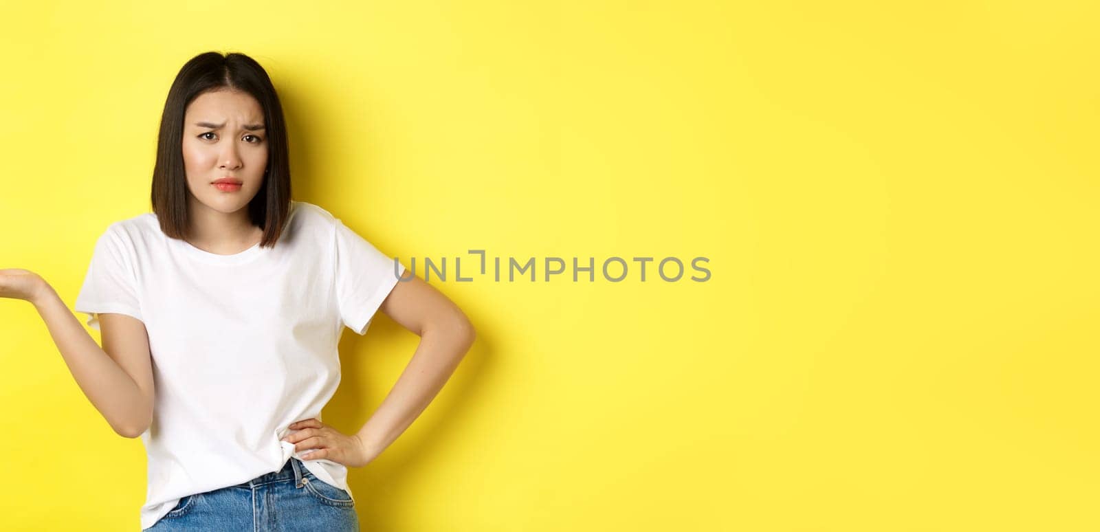 Disappointed asian woman asking so what, raise hand up and staring skeptical at camera, standing over yellow background.