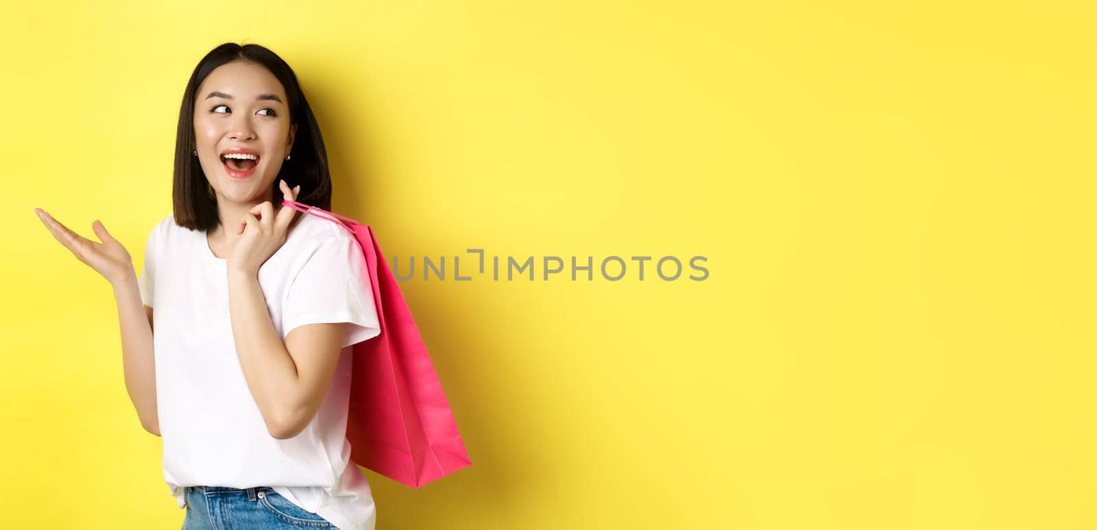 Cheerful asian female shopper looking amused, holding shopping bag and looking right, standing over yellow background.