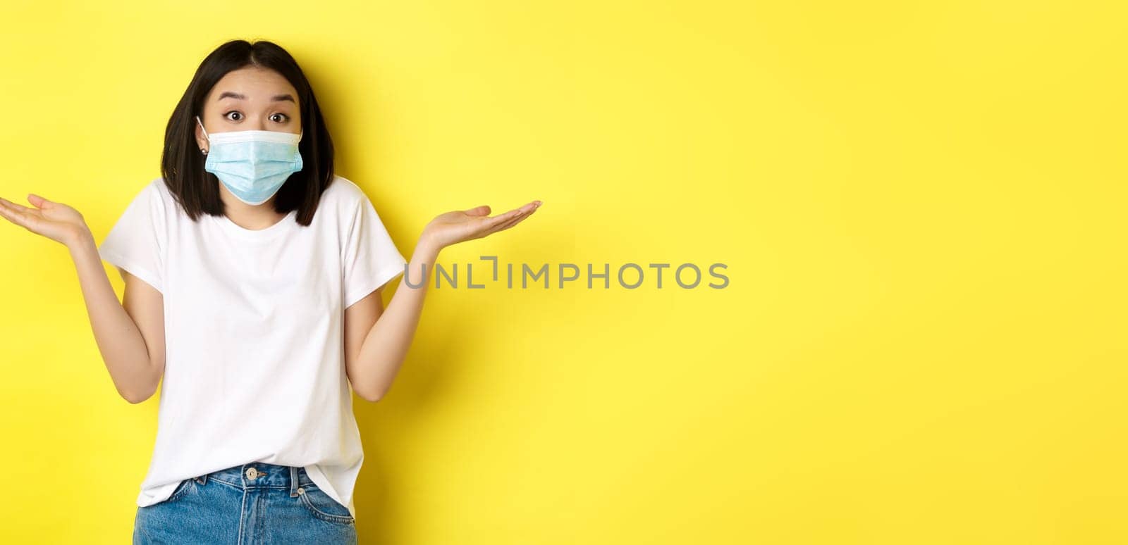 Covid-19, quarantine and social distancing concept. Clueless asian woman in medical mask shrugging shoulders, spread hands sideways clueless, know nothing, yellow background by Benzoix