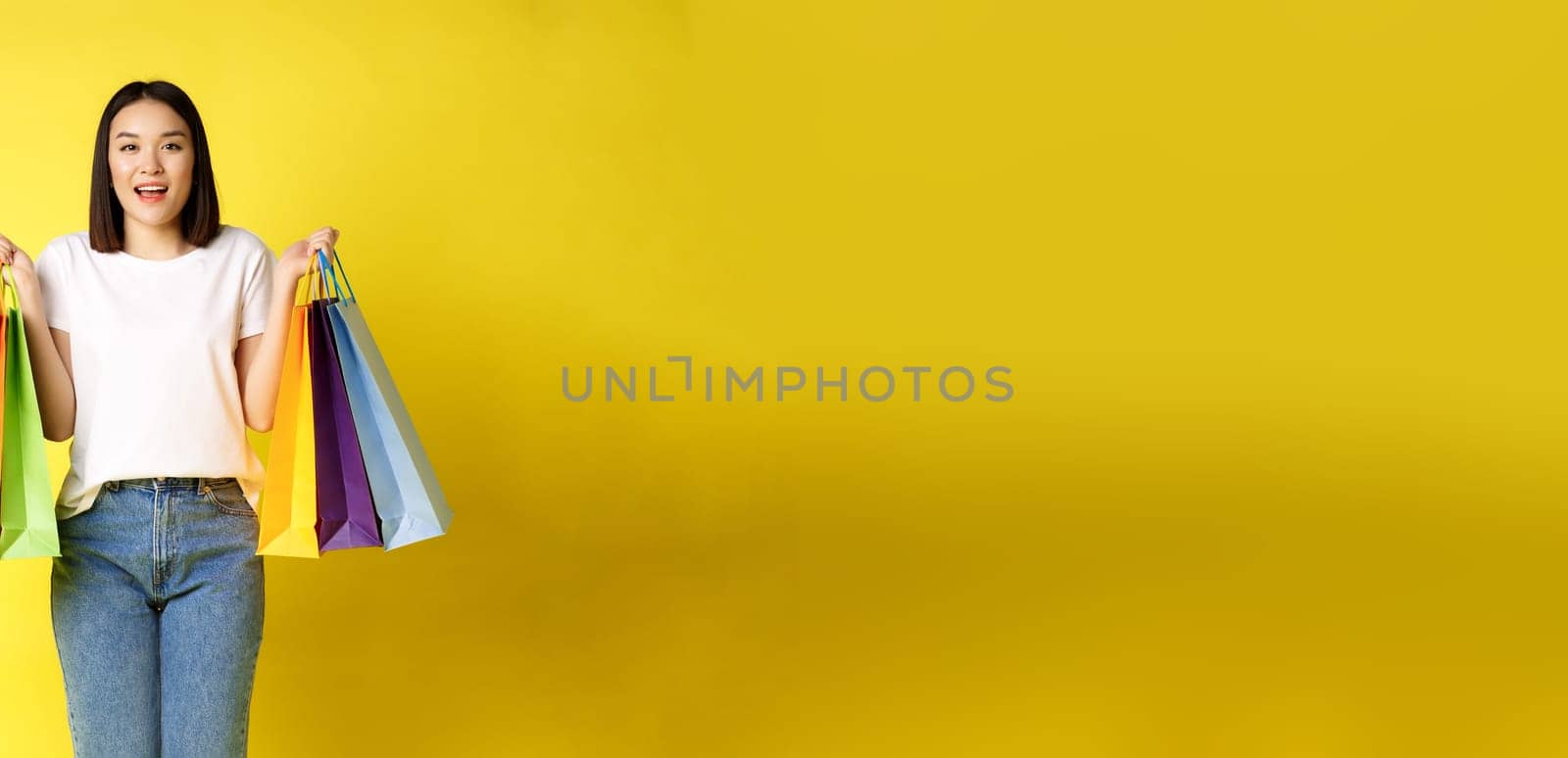 Full size portrait of beautiful asian girl going shopping, holding paper bags from stores and smiling, standing in jeans and white t-shirt over yellow background.