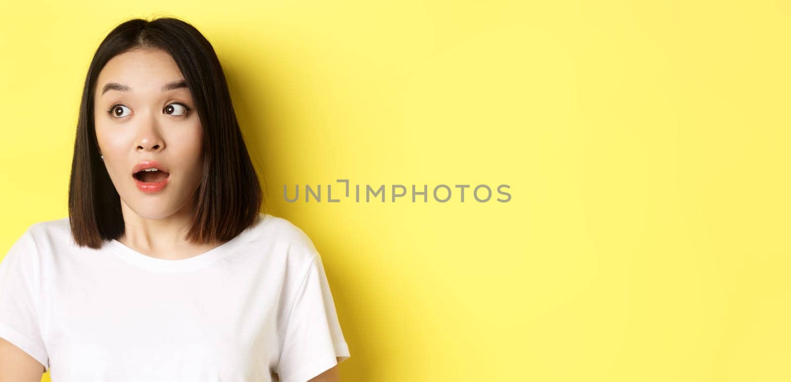 Close up of surprised asian girl drop jaw, gasping and looking left at logo, standing over yellow background by Benzoix