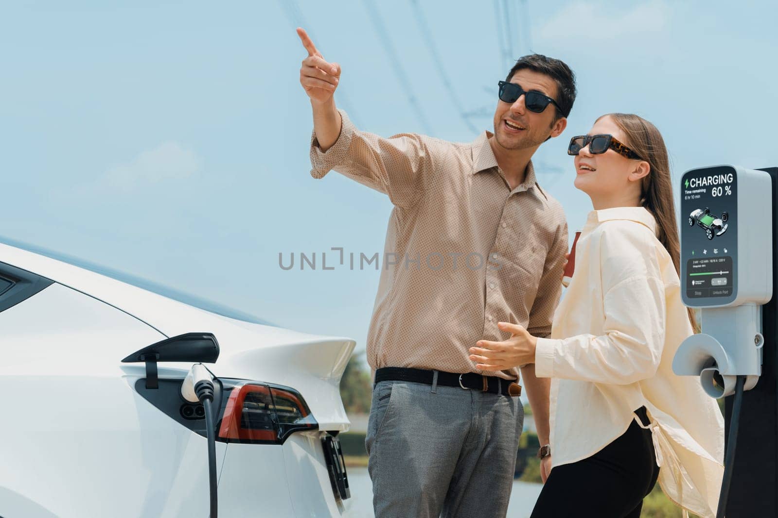 Young couple recharge electric car's battery from charging station in outdoor green city park in springtime. Rechargeable EV car for sustainable environmental friendly urban travel lifestyle.Expedient