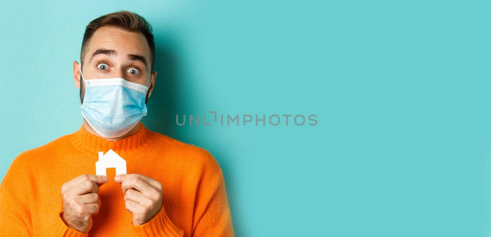 Real estate and coronavirus pandemic concept. Close-up of adult man in medical mask holding small paper house maket and smiling, searching for apartment, light blue background by Benzoix