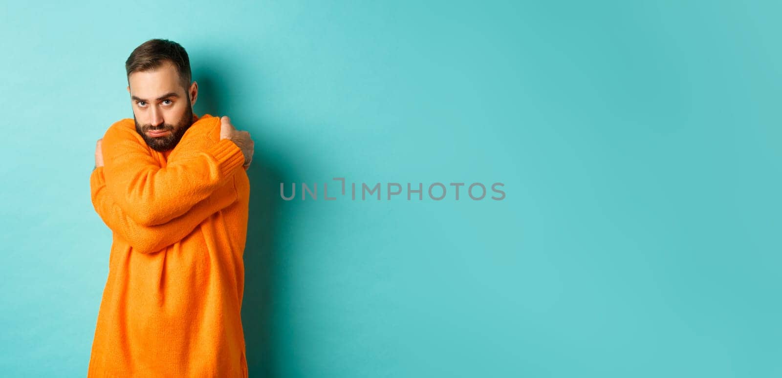 Timid man feeling offended and defensive, hugging himself and looking suspicious at camera, standing over light blue background.