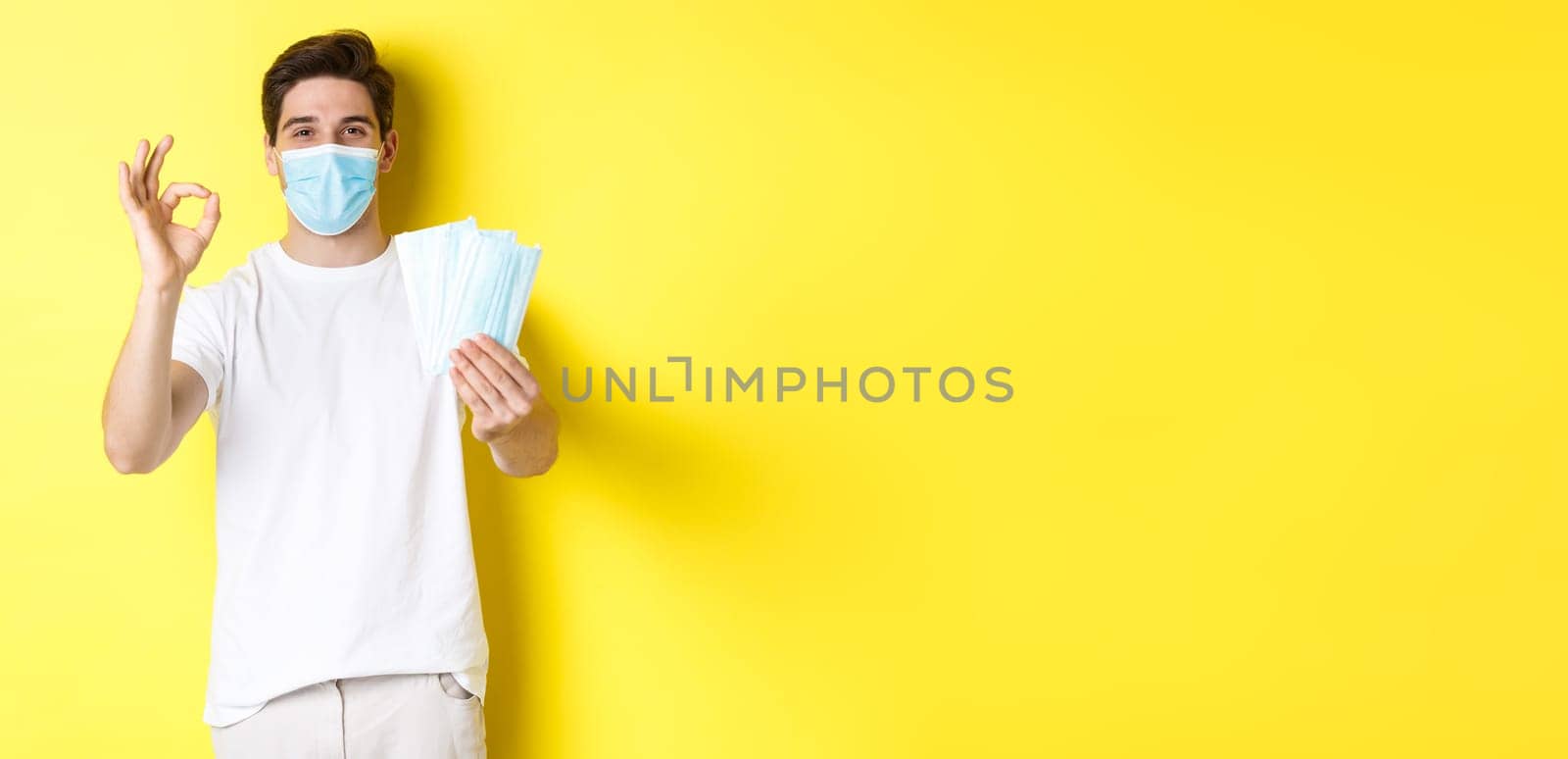 Concept of covid-19, quarantine and preventive measures. Satisfied man showing okay sign and giving medical masks, standing over yellow background.