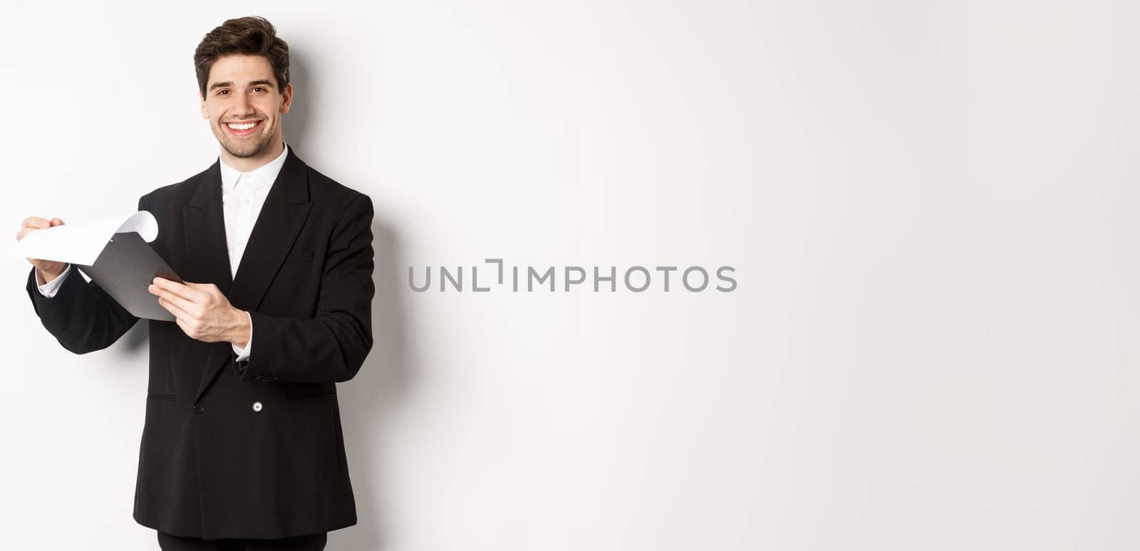 Image of confident businessman in suit looking at documents, holding clipboard and smiling pleased, standing against white background.