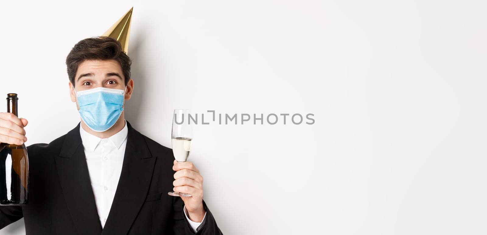 Concept of party during covid-19. Close-up of handsome man in suit, funny hat and medical mask, holding bottle of champagne, celebrating new year during coronavirus by Benzoix