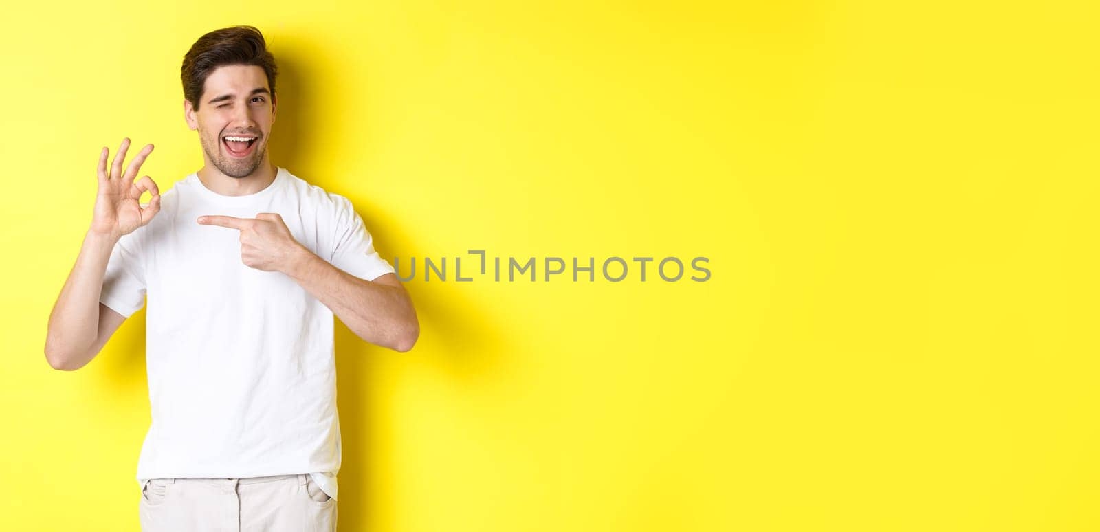 Image of handsome young man approve something, showing okay sign and winking, standing against yellow background.