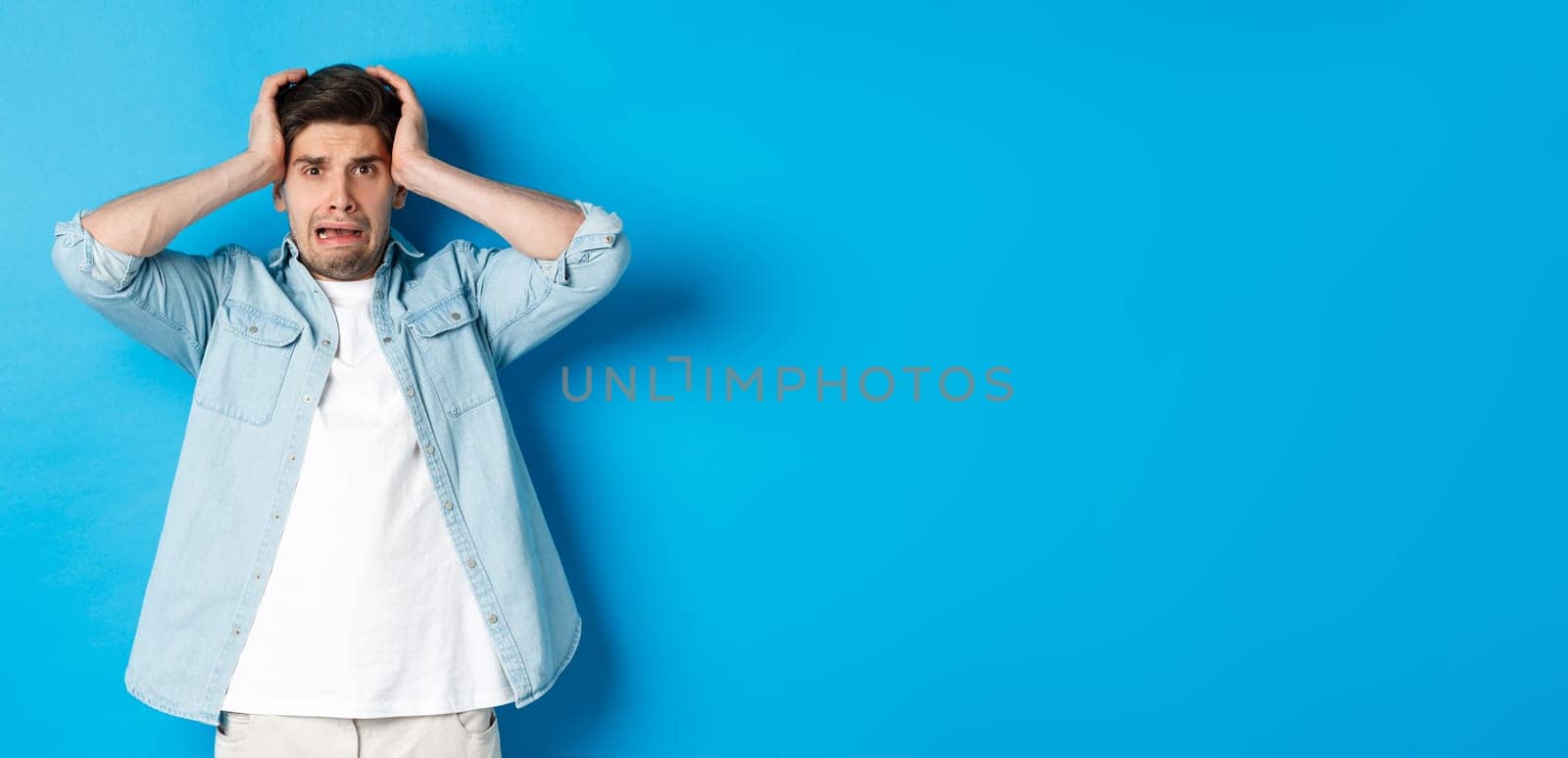 Image of man in panic holding hands on head, looking frustrated and anxious, standing against blue background by Benzoix