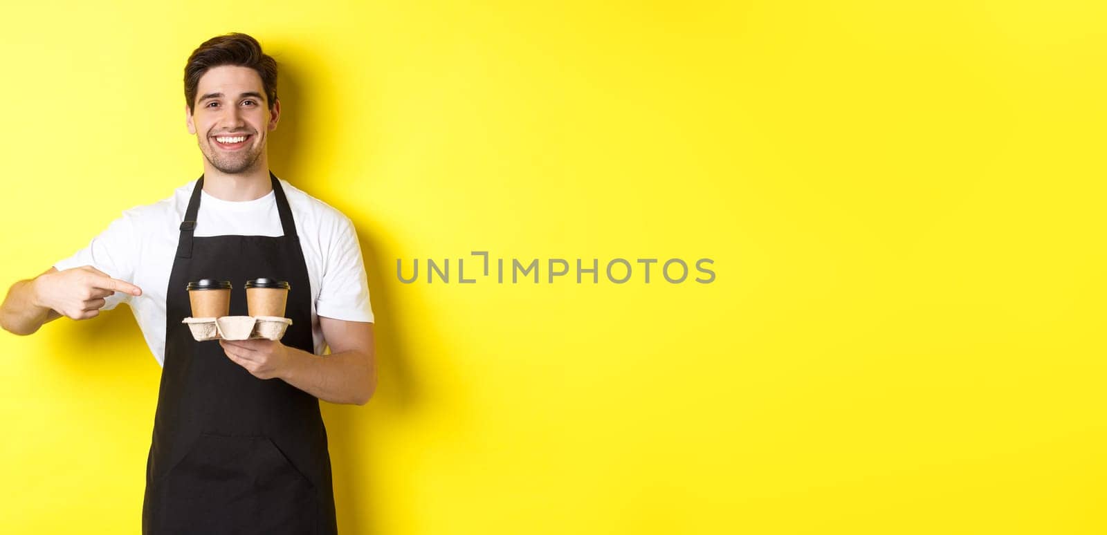 Handsome barista holding two cups of takeaway coffee, pointing finger at drinks and smiling, standing in black apron against yellow background.