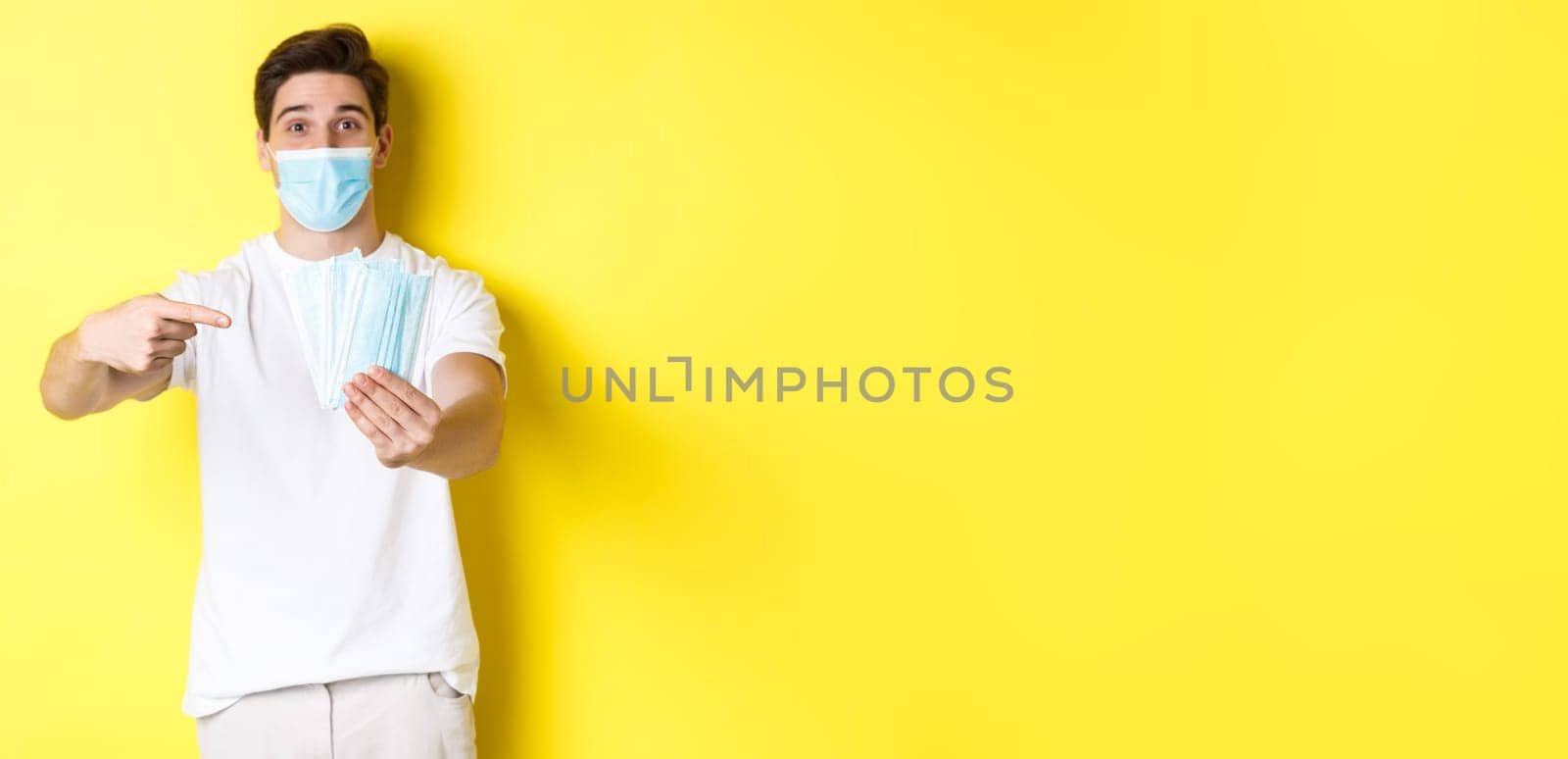 Concept of covid-19, quarantine and preventive measures. Young caucasian man giving medical masks for you, standing against yellow background by Benzoix